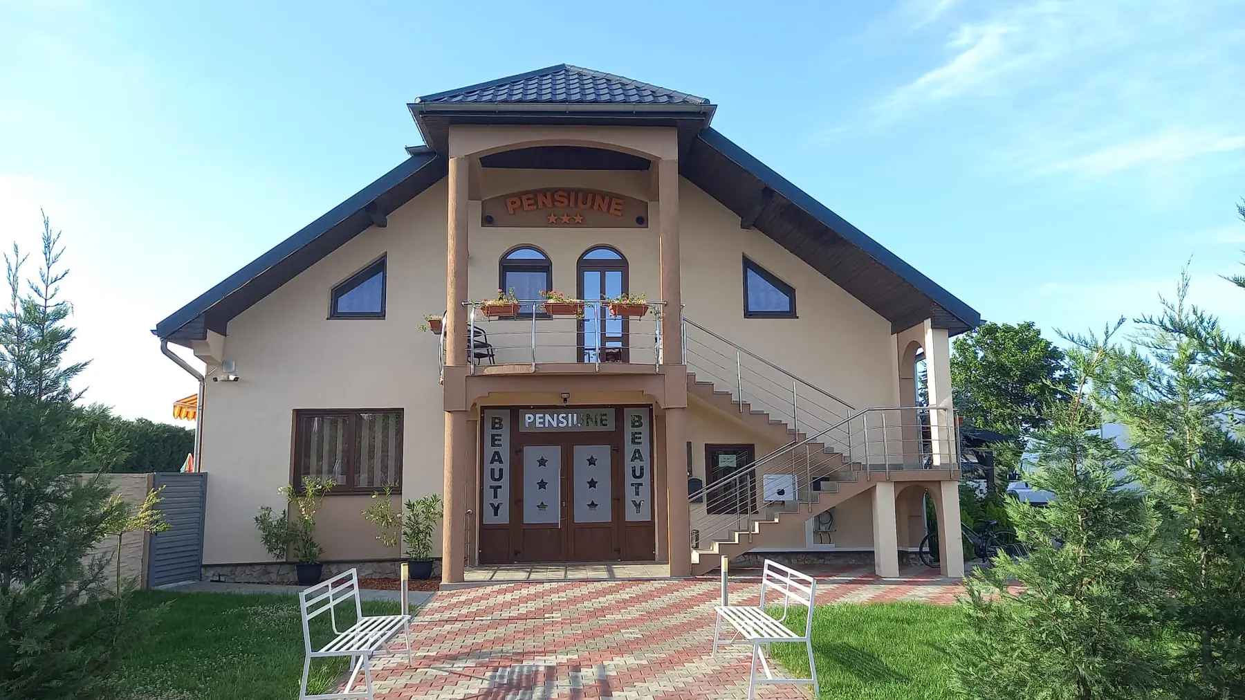 Fațada Pensiunii Beauty, cu firmă luminoasă, bănci albe și grădină, într-o dimineață senină