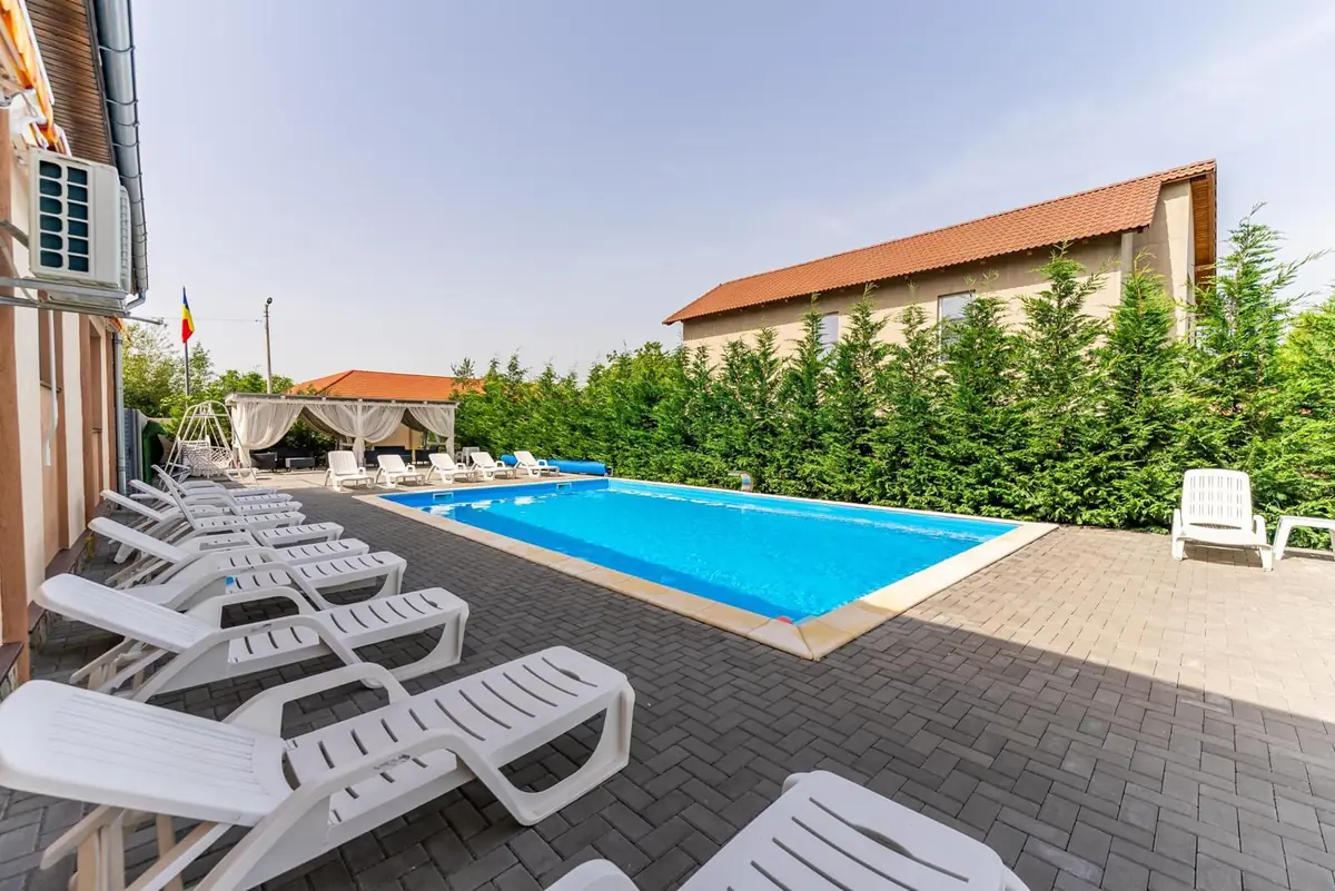 Piscină dreptunghiulară cu apă albastră, șezlonguri albe și gard viu din tuia